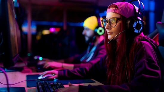Eine junge Erwachsene mit Gamingbrille und Headset sitzt vor einem Bildschirm und spielt Computerspiele. Im Hintergrund sitzt ein junger Erwachsener mit Kopfhörern auf