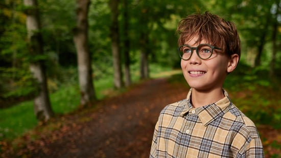 Ein Junge im karierten Hemd und mit einer Brille steht lächelnd im Wald