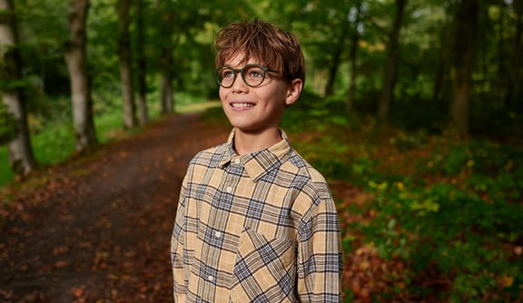 Ein Junge im karierten Hemd und mit einer Brille steht lächelnd im Wald