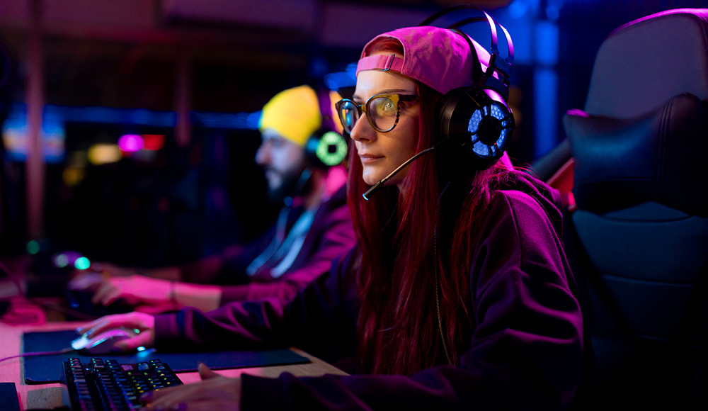 Eine junge Erwachsene mit Gamingbrille und Headset sitzt vor einem Bildschirm und spielt Computerspiele. Im Hintergrund sitzt ein junger Erwachsener mit Kopfhörern auf