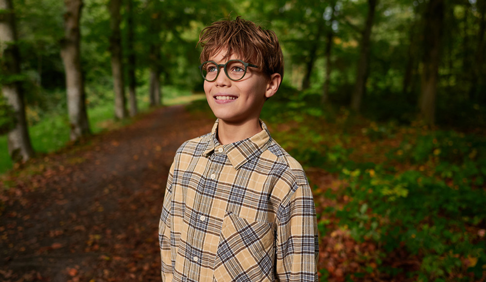 Ein Junge im karierten Hemd und mit einer Brille steht lächelnd im Wald